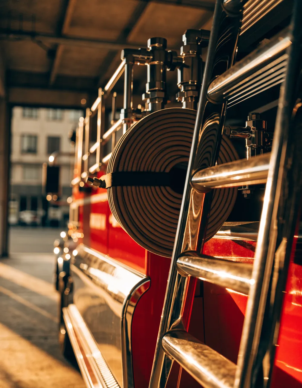 Polished chrome, coiled hose and ladder rungs on a red fire engine in a station bay at golden hour